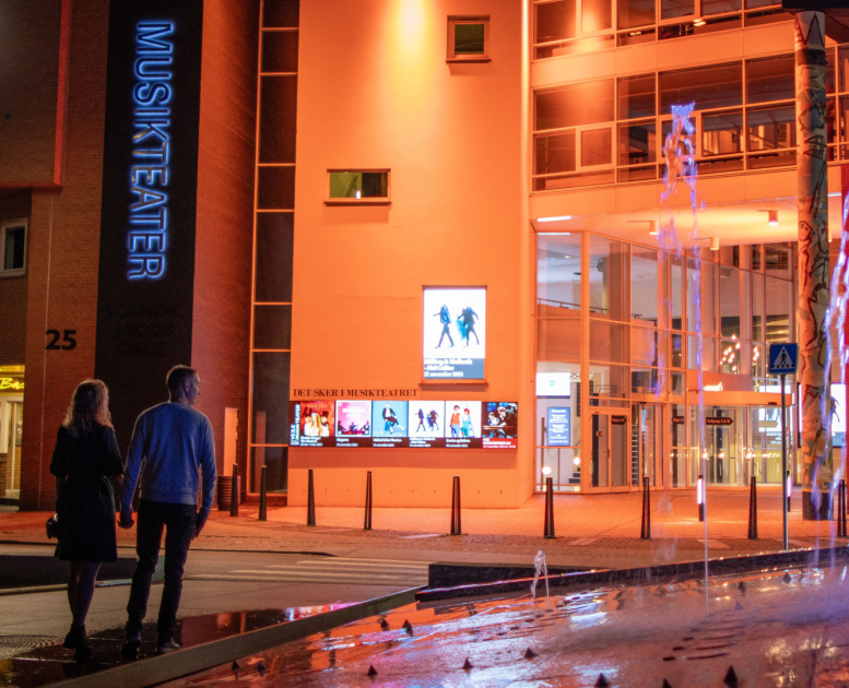 A couple on their way to Vejle Musikteater walks across the roundabout with the fountain out in front of the music theatre